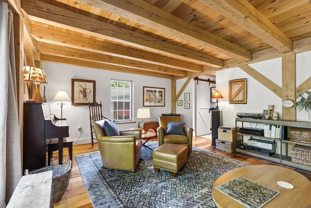 This cozy living room features exposed timber ceiling beams and warm wood flooring, creating a rustic and inviting atmosphere. The space is furnished with two leather armchairs, a matching ottoman, and a piano, all arranged on a large patterned area rug. A metal shelving unit provides storage for books and media, while a barn-style sliding door adds a touch of farmhouse charm to the room.