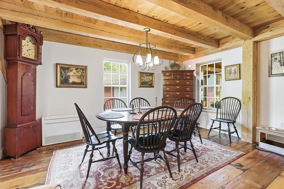 This rustic dining room features exposed wooden ceiling beams and warm wood flooring, creating a cozy, traditional atmosphere. A dark wood dining table is surrounded by matching spindle-back chairs, centered beneath a classic chandelier, while a tall grandfather clock stands prominently in the corner. The space is accented by a patterned area rug, framed artwork on the walls, and a tall wooden chest of drawers, all illuminated by natural light from the windows.
