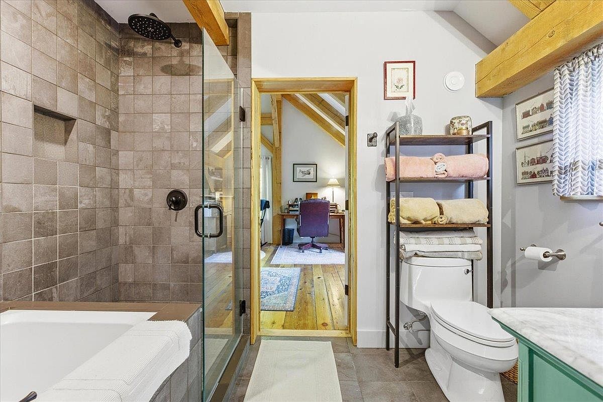 This bathroom features a walk-in shower with tiled walls and a glass door, positioned next to a bathtub. A metal shelving unit sits above the toilet, providing storage for towels, while a doorway leads directly into an adjacent office space with wood flooring. The room incorporates rustic elements like exposed wooden beams, creating a warm and functional transition between living areas.