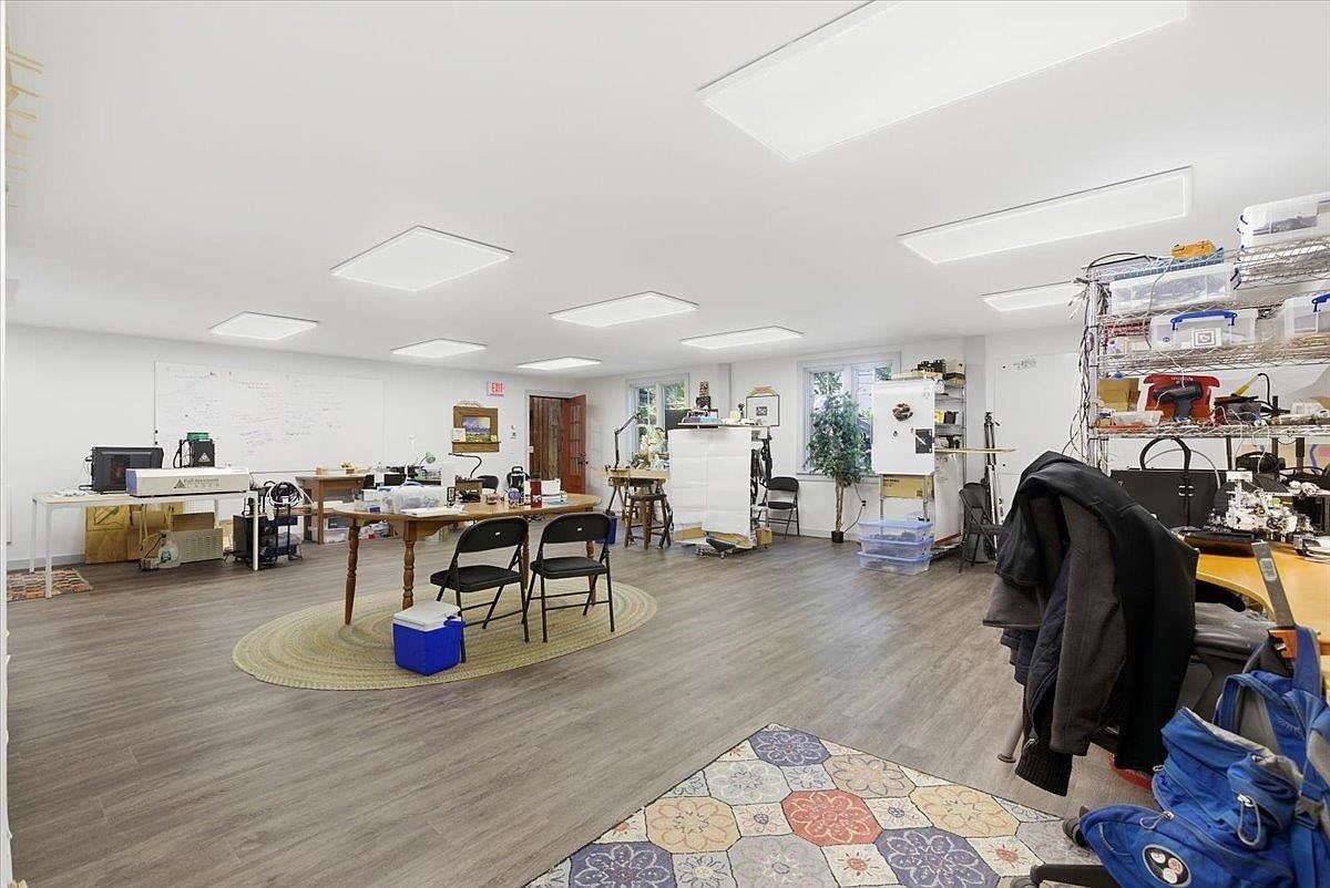 This spacious, multi-purpose room functions as a bright and functional workspace or hobby studio. The area features light-toned wood-look flooring, recessed ceiling lighting, and a large whiteboard on the wall, providing an open layout suitable for collaborative projects or creative endeavors. Multiple tables, chairs, and storage shelving units are arranged throughout the room, creating a versatile environment that could easily be adapted for various professional or personal uses.