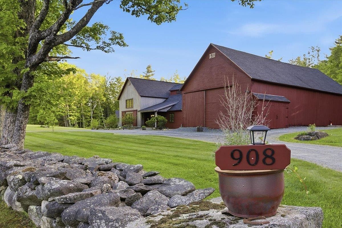 This picturesque property features a classic red barn-style structure paired with a modern farmhouse addition, set against a lush green lawn. A rustic stone wall runs along the foreground, leading the eye toward a decorative planter displaying the address '908'. The scene is framed by mature trees under a clear blue sky, evoking a serene and well-maintained country estate atmosphere.