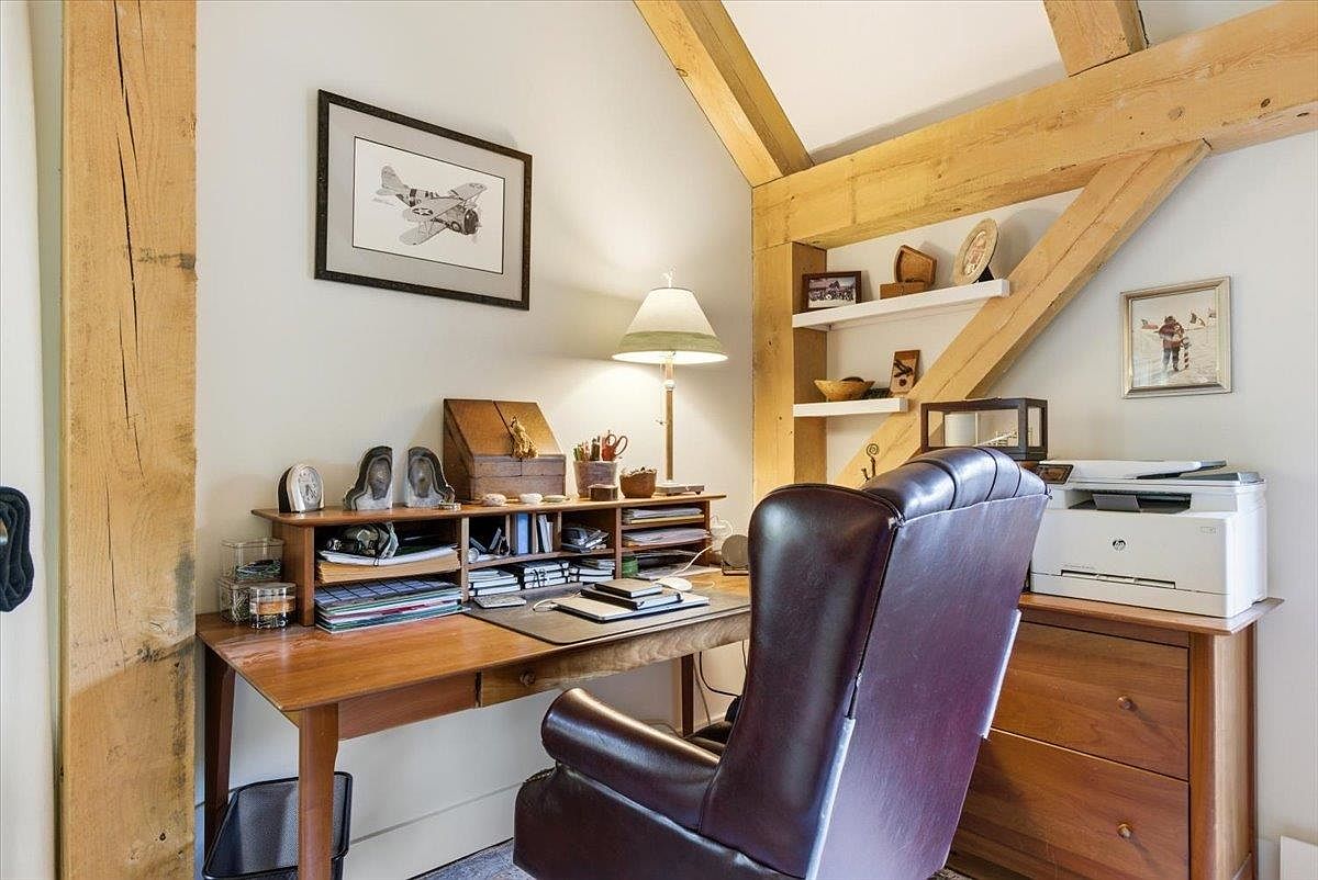 This cozy home office features a warm, rustic aesthetic with exposed timber beams and a sloped ceiling. A wooden desk is organized with various office supplies and a lamp, paired with a comfortable leather chair, while built-in shelving displays decorative items. The space is well-lit and includes a printer on a matching wooden cabinet, creating a functional and inviting workspace.
