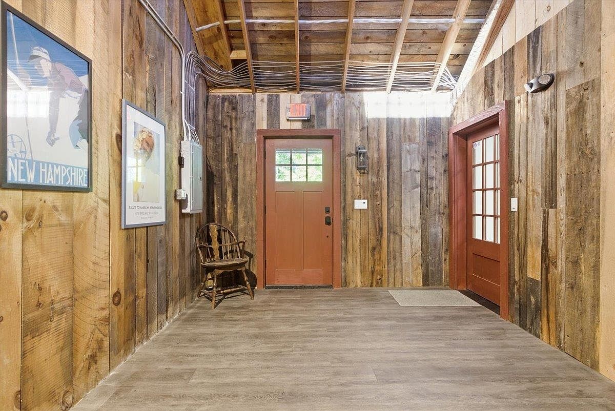 This rustic entryway features reclaimed wood plank walls and a neutral wood-look floor, creating a warm and textured aesthetic. The space includes a wooden chair, two framed posters on the wall, and two distinct doors, one of which is a glass-paneled door leading to the exterior. The exposed ceiling rafters and visible electrical conduit add to the industrial-farmhouse character of the room.