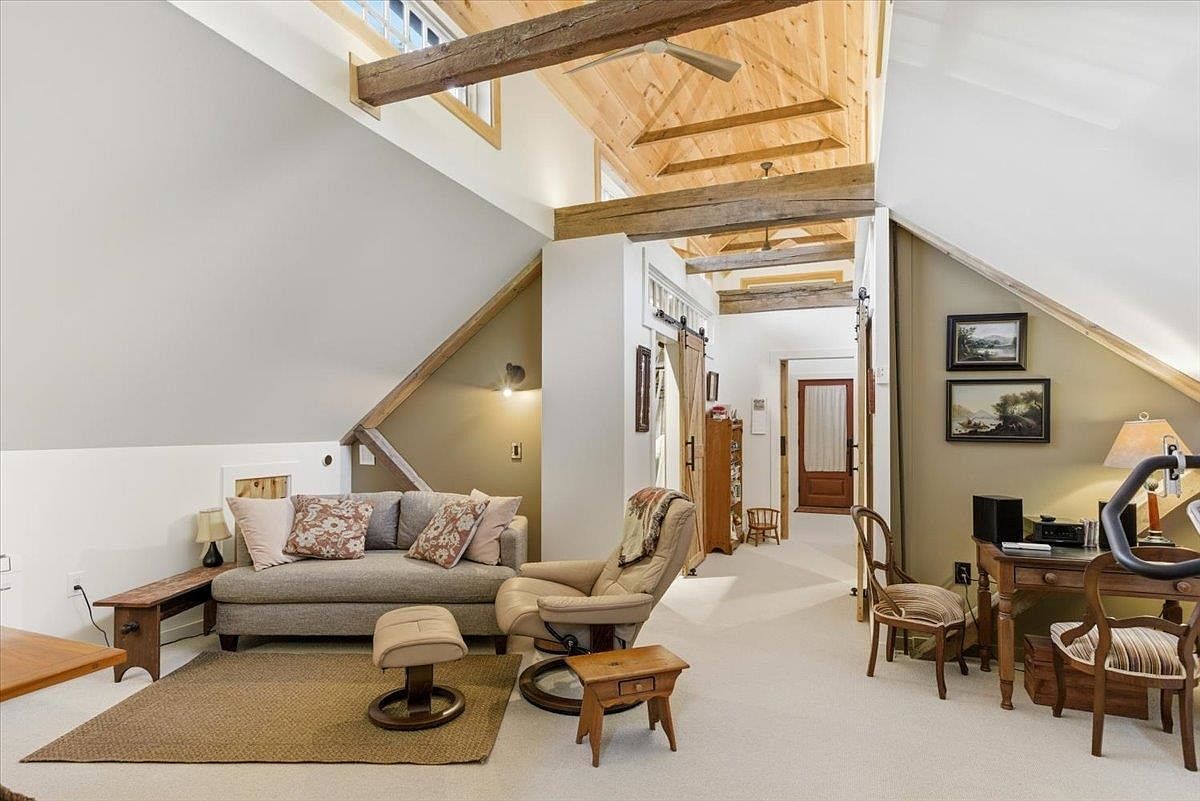 This spacious attic living area features a vaulted ceiling with exposed wooden beams and a skylight that fills the room with natural light. The space is furnished with a comfortable sofa, a recliner, and a wooden desk, creating a cozy multi-purpose environment. The room is characterized by its warm, rustic aesthetic, highlighted by the angled walls and neutral carpeting.