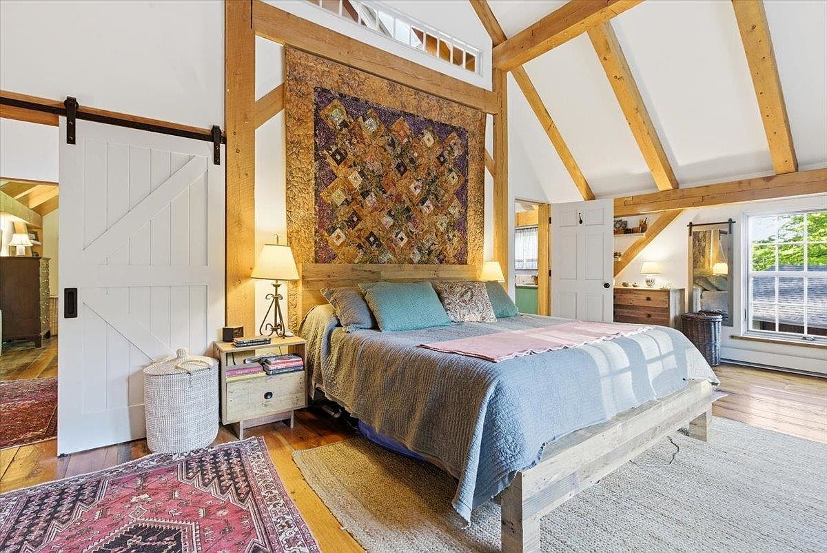 This rustic-inspired bedroom features exposed timber beams and a vaulted ceiling, creating an airy and warm atmosphere. A large, colorful quilt serves as a focal point above the bed, which is dressed in neutral linens and accented with teal pillows. The room includes a sliding barn door, light wood furniture, and a mix of patterned area rugs, all bathed in natural light from the windows.