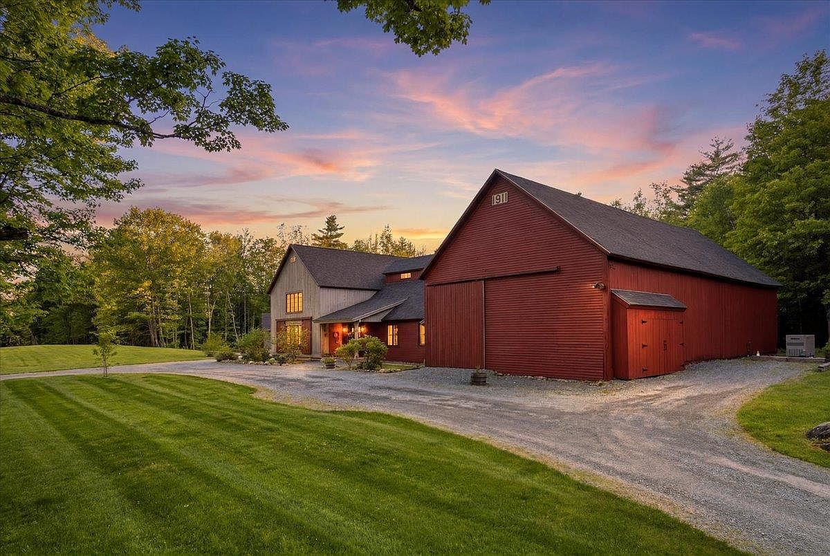This picturesque property features a charming red barn-style structure connected to a multi-level home with grey siding, set against a vibrant sunset sky. A gravel driveway leads up to the buildings, framed by a well-manicured green lawn and mature trees. The scene captures a serene, rustic aesthetic with a warm, inviting atmosphere perfect for a country estate.