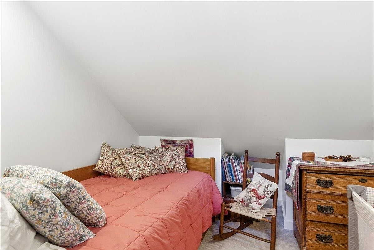 This cozy bedroom is tucked under a sloped ceiling, creating an intimate and inviting atmosphere. The room features a twin bed with a coral-colored quilt, a wooden rocking chair, and a vintage-style wooden dresser. A small bookshelf sits beside the bed, and the space is accented with patterned throw pillows, offering a charming and rustic aesthetic.