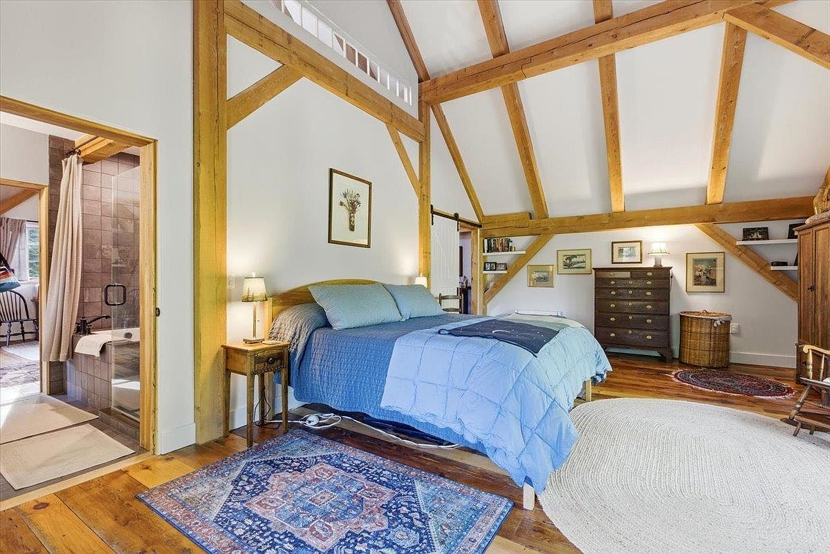 This bedroom features a rustic aesthetic with exposed timber beams and vaulted ceilings, creating an airy and open atmosphere. The room is furnished with a bed dressed in blue bedding, a wooden nightstand, and a dark wood dresser, all set against a backdrop of light-colored walls and warm wood flooring. A doorway to the left reveals a tiled bathroom, while a patterned area rug and a circular white rug add texture to the space.