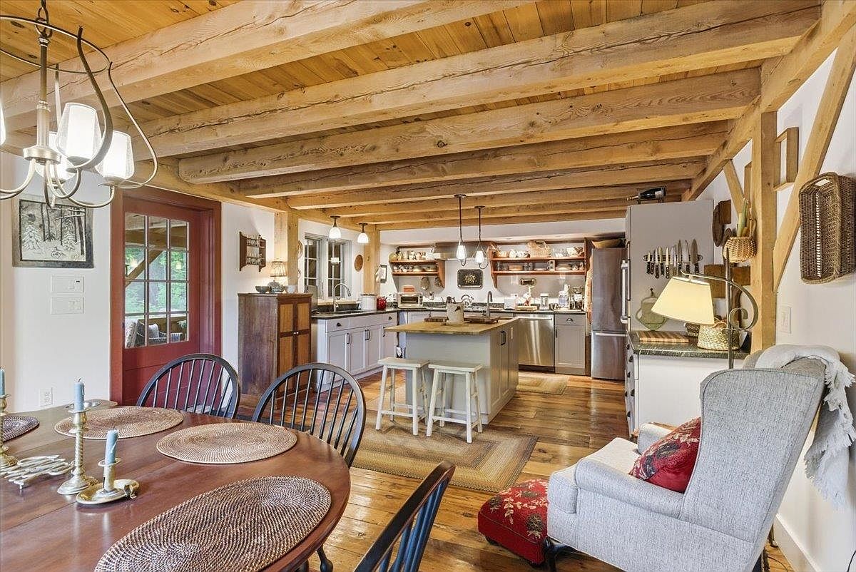 This open-concept space features a rustic kitchen with exposed wooden ceiling beams and warm wood flooring, seamlessly flowing into a dining area. The kitchen is equipped with light-toned cabinetry, a central island with bar seating, and stainless steel appliances, while a cozy armchair sits in the foreground. The perspective captures the inviting, farmhouse-inspired aesthetic of the home, highlighting the warm textures and functional layout.