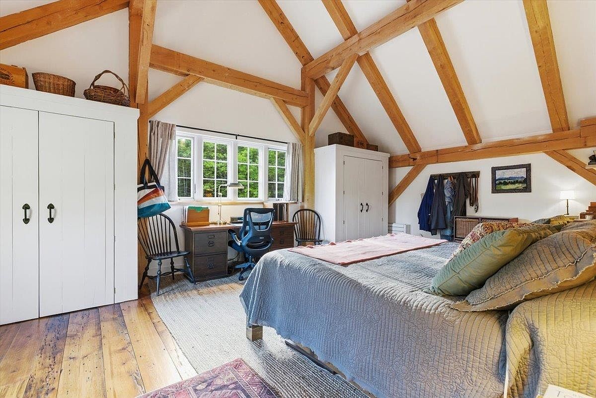 This charming bedroom features exposed timber beams and a vaulted ceiling, creating a rustic and airy atmosphere. The room is furnished with a comfortable bed, a white wardrobe, and a dedicated workspace positioned under a set of windows that bring in natural light. The warm wood flooring and neutral color palette contribute to a cozy, inviting aesthetic suitable for a guest room or home office combination.