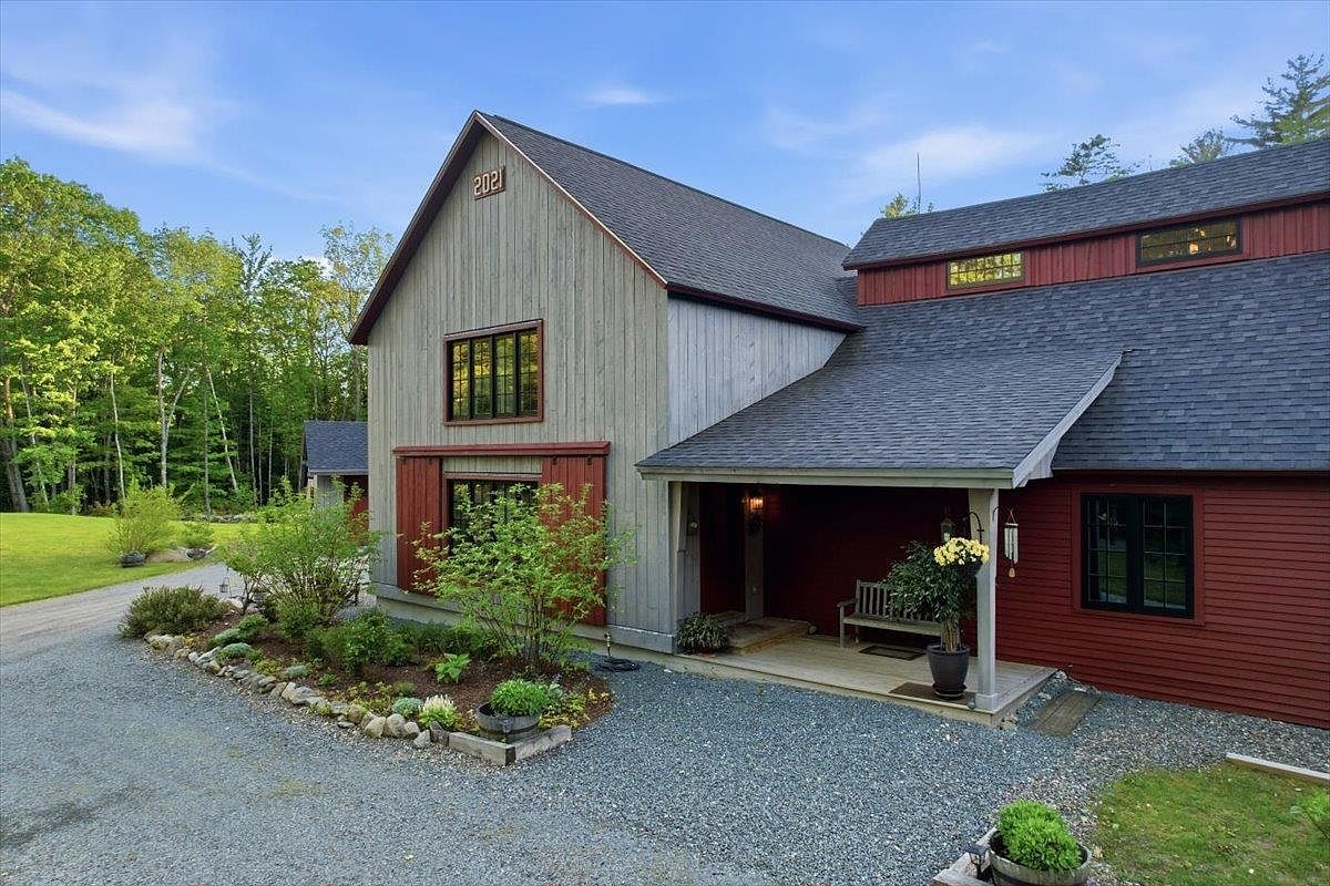 This charming home features a blend of rustic and modern farmhouse aesthetics, highlighted by vertical gray siding and contrasting red accents. A covered porch with a wooden bench provides a welcoming entryway, while the gravel driveway leads up to the structure. The exterior is accented by dark-framed windows and a prominent '2021' date plaque on the gable, set against a backdrop of lush green trees.