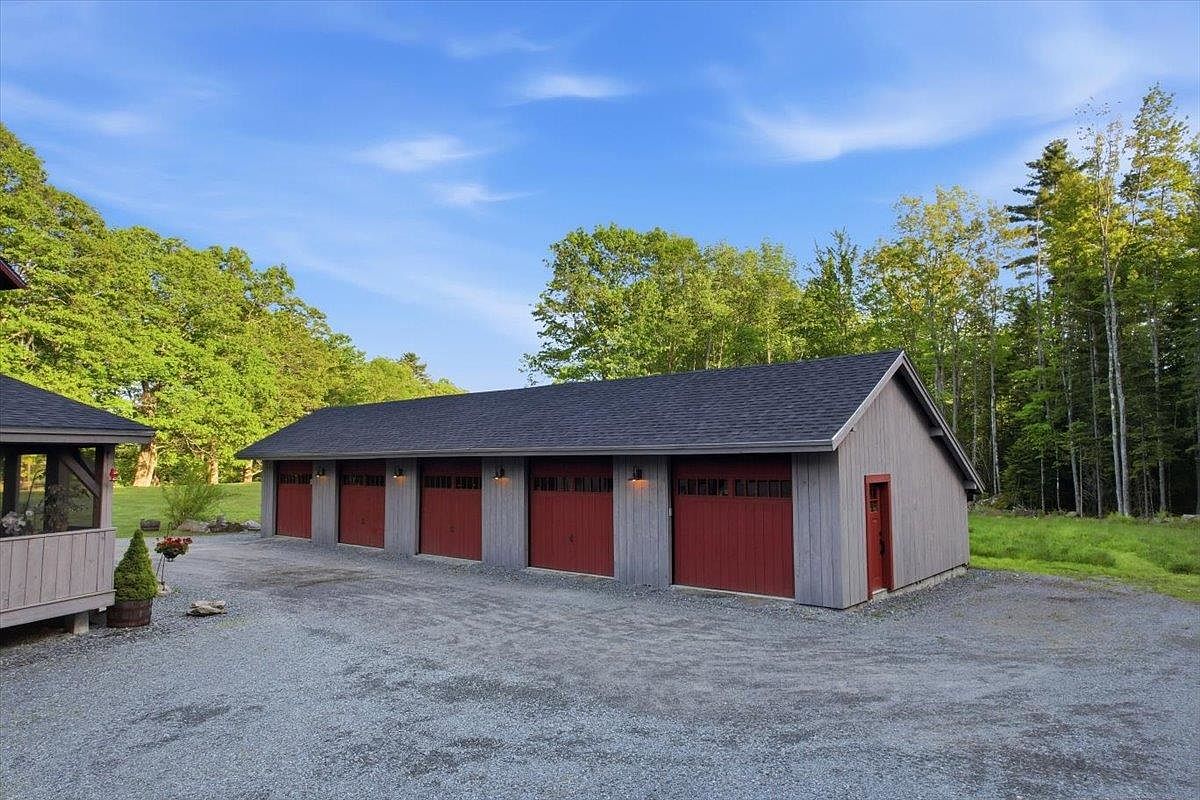 This image showcases a spacious, detached five-bay garage with a classic, rustic aesthetic. The structure features vertical wood siding painted in a neutral grey tone, contrasted by vibrant red garage doors and a matching side entry door. Set against a backdrop of lush green trees and a clear blue sky, the gravel driveway leads up to the building, emphasizing its functional and well-maintained appearance.