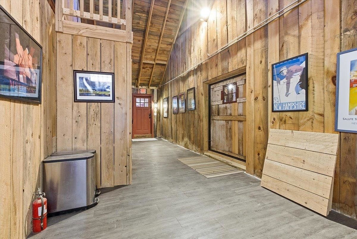 This rustic-style hallway features warm, vertical wood-plank walls and a neutral wood-look floor. The space includes a red door at the end of the corridor, a metal trash receptacle, and several framed posters adorning the walls. The perspective is a straight-on shot looking down the hall, highlighting the textured wood surfaces and the transition toward the entryway.