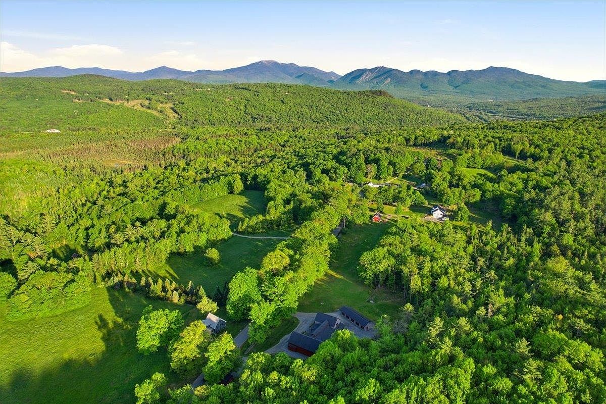 This high-angle aerial perspective captures a sprawling rural property nestled within a lush, green landscape. The scene features a primary residence with a dark roof and red siding, surrounded by expansive lawns, mature trees, and a distant mountain range under a clear blue sky. The composition emphasizes the property's secluded, natural setting and its integration into the surrounding forest.