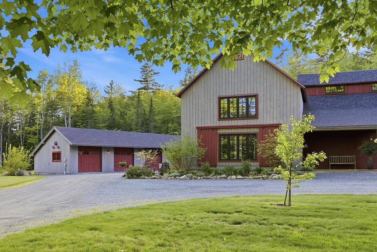 This charming rural property features a large, barn-style home with vertical gray siding and contrasting red trim around the windows and garage doors. A gravel driveway leads up to the attached multi-bay garage, while a lush green lawn and mature trees frame the scene under a clear blue sky. The architecture blends rustic barn aesthetics with modern residential comfort, creating a welcoming and serene atmosphere.