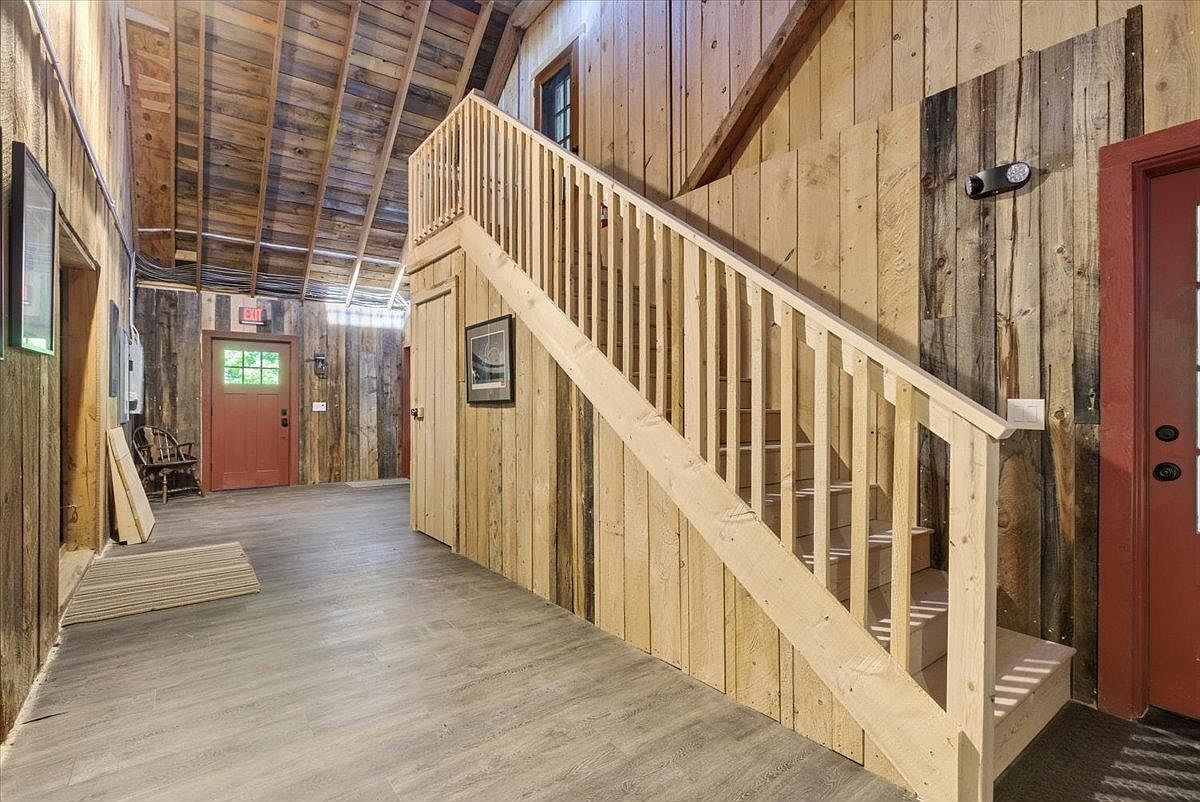 This interior space features a rustic aesthetic with vertical wood-plank walls and a prominent wooden staircase leading to an upper level. The area includes a light-toned wood-look floor, a red door with glass panes, and an open-rafter ceiling that adds to the barn-like character of the home. The perspective is from the ground floor, looking toward the staircase and the exit door, highlighting the open layout and architectural woodwork.
