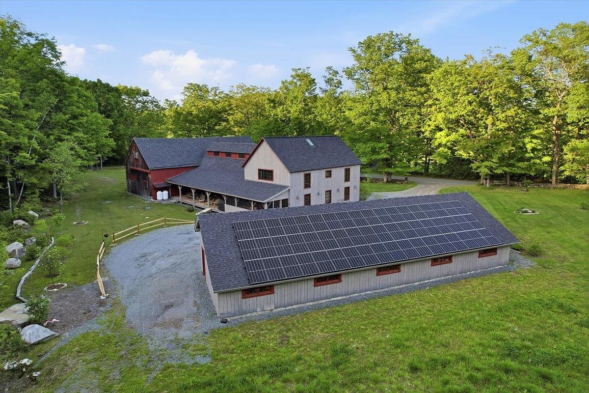 This aerial perspective captures a sprawling rural property featuring a complex of connected barn-style buildings with a mix of red and grey siding. A prominent, long outbuilding in the foreground is equipped with a large array of solar panels on its roof, set against a backdrop of lush green trees and a gravel driveway. The scene conveys a peaceful, sustainable country lifestyle with ample open space and mature landscaping.