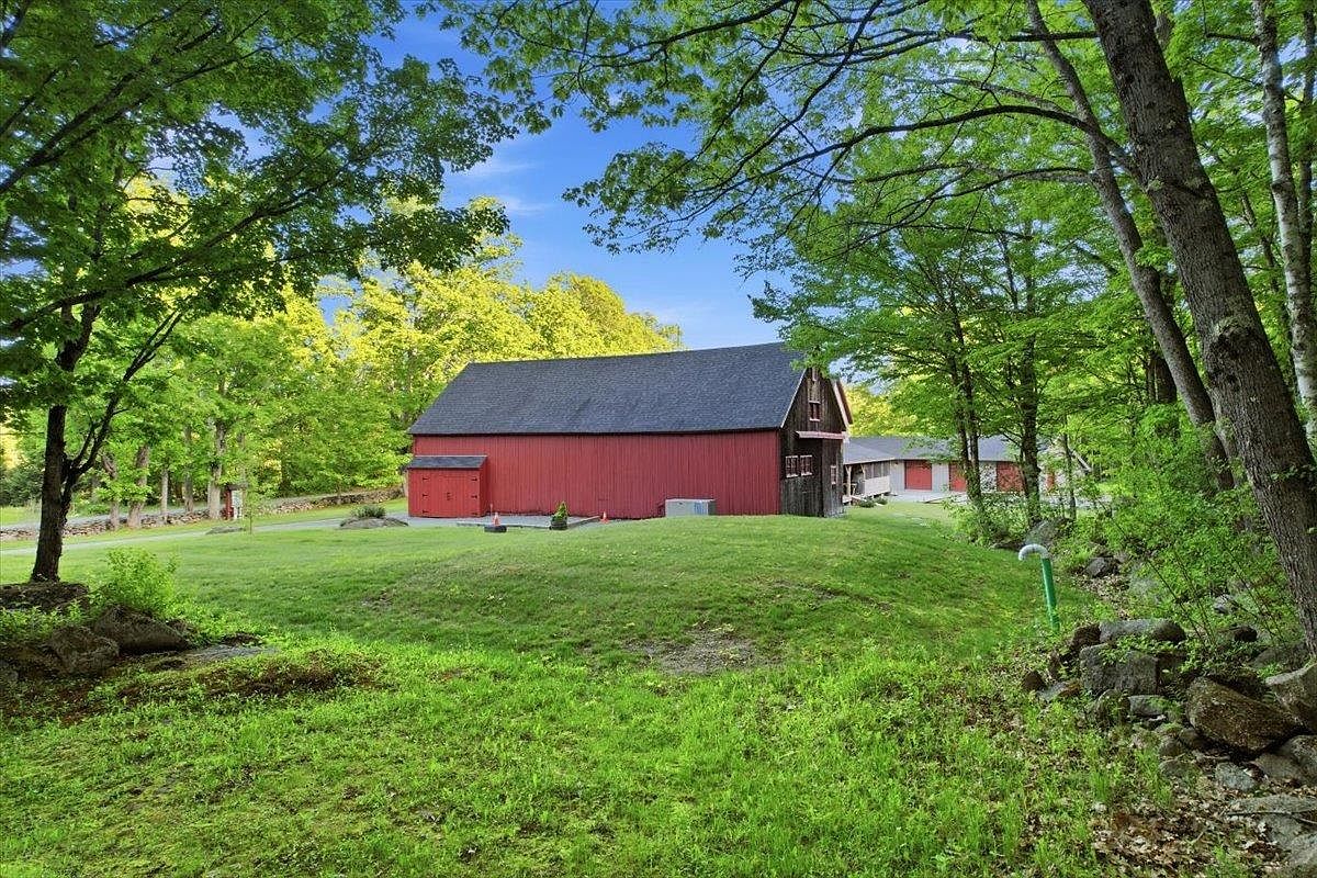 This image captures a side view of a classic red barn set against a lush, green landscape. The structure features a dark shingled roof and a prominent side entrance, with a secondary building visible in the background. The scene is framed by mature trees, creating a peaceful and rustic atmosphere typical of a rural property.