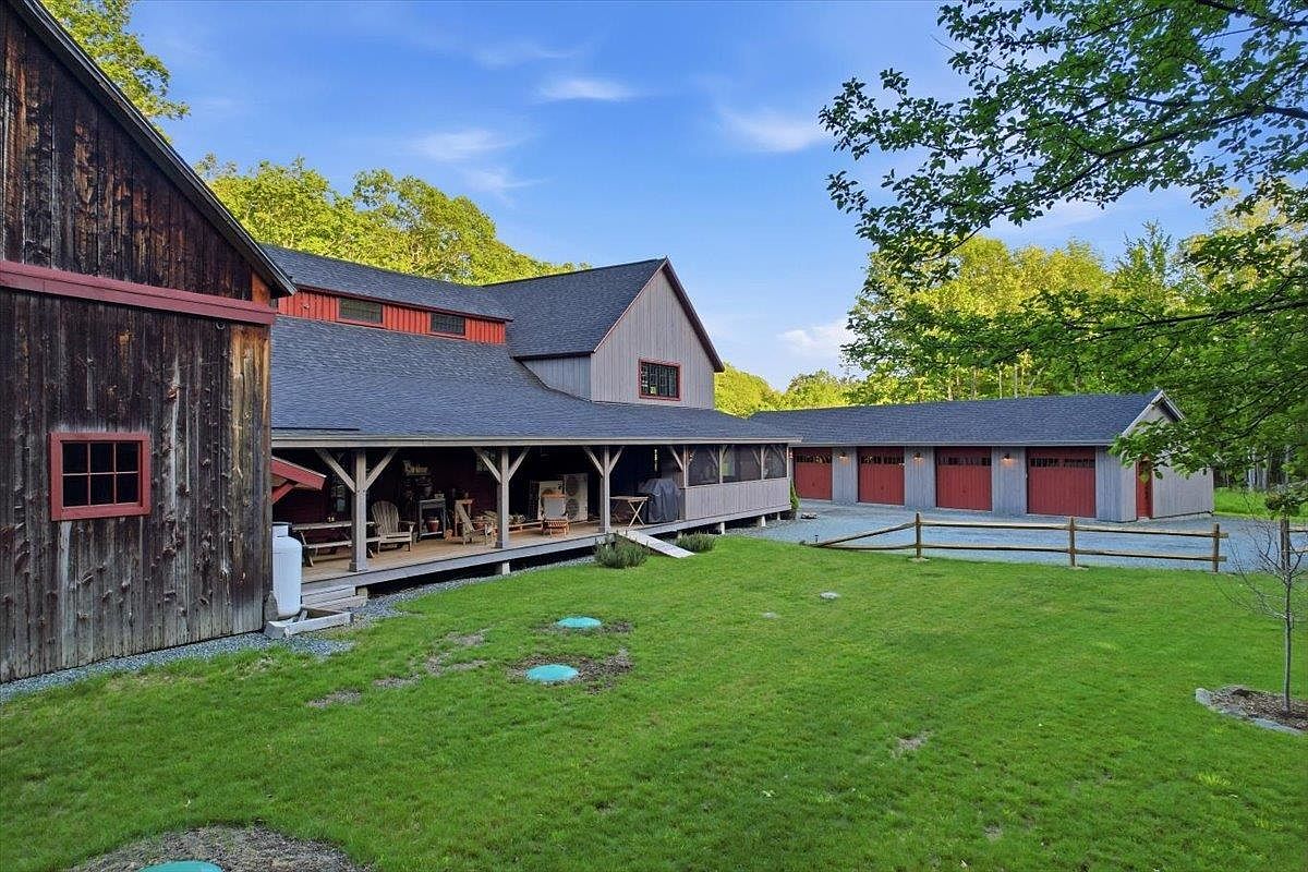 This rear view of a rustic-style home showcases a sprawling covered porch that spans the length of the main structure, offering a shaded outdoor living space. The property features a mix of weathered wood siding and painted grey accents, complemented by a detached multi-bay garage with matching red doors. A well-maintained lawn stretches out toward a simple wooden fence, creating a peaceful and expansive backyard setting.