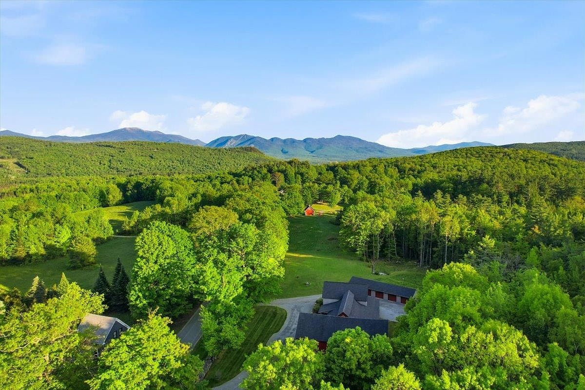 This high-angle aerial view captures a sprawling rural property nestled within a lush, green landscape surrounded by rolling mountains. The scene features a main residence with a dark roof, a paved driveway, and a small red barn situated in the middle of a large, open meadow. The expansive forest and clear blue sky create a serene and private atmosphere, highlighting the property's secluded natural setting.