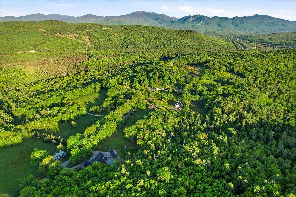 This high-altitude aerial perspective captures a sprawling, lush property nestled within a dense forest landscape. The scene features multiple residential structures surrounded by expansive green lawns and winding pathways, all set against a backdrop of rolling mountain ranges. The composition emphasizes the property's secluded, natural setting and its integration into the surrounding woodland.