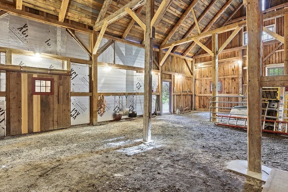 This image captures the interior of a rustic, open-concept barn featuring exposed timber framing and a dirt floor. The space is illuminated by overhead lighting and natural light filtering through small windows, with silver insulation panels lining one wall. A metal gate and a ladder are visible in the background, emphasizing the functional, agricultural nature of the structure.
