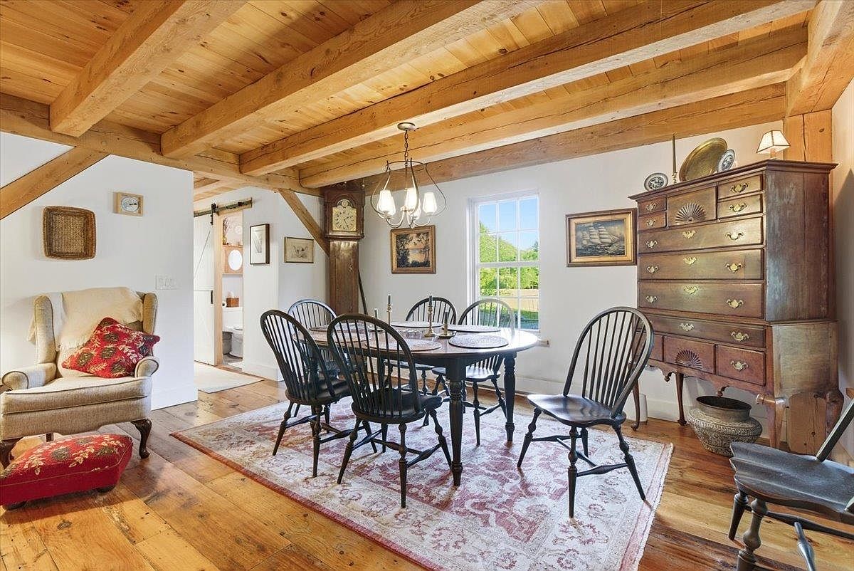 This rustic dining room features exposed wooden ceiling beams and warm wood flooring, creating a cozy, traditional atmosphere. The space is furnished with a dark wood dining table surrounded by spindle-back chairs, a tall grandfather clock, and a large antique-style wooden chest of drawers. A patterned area rug anchors the dining area, while a window provides natural light and a view of the outdoors.