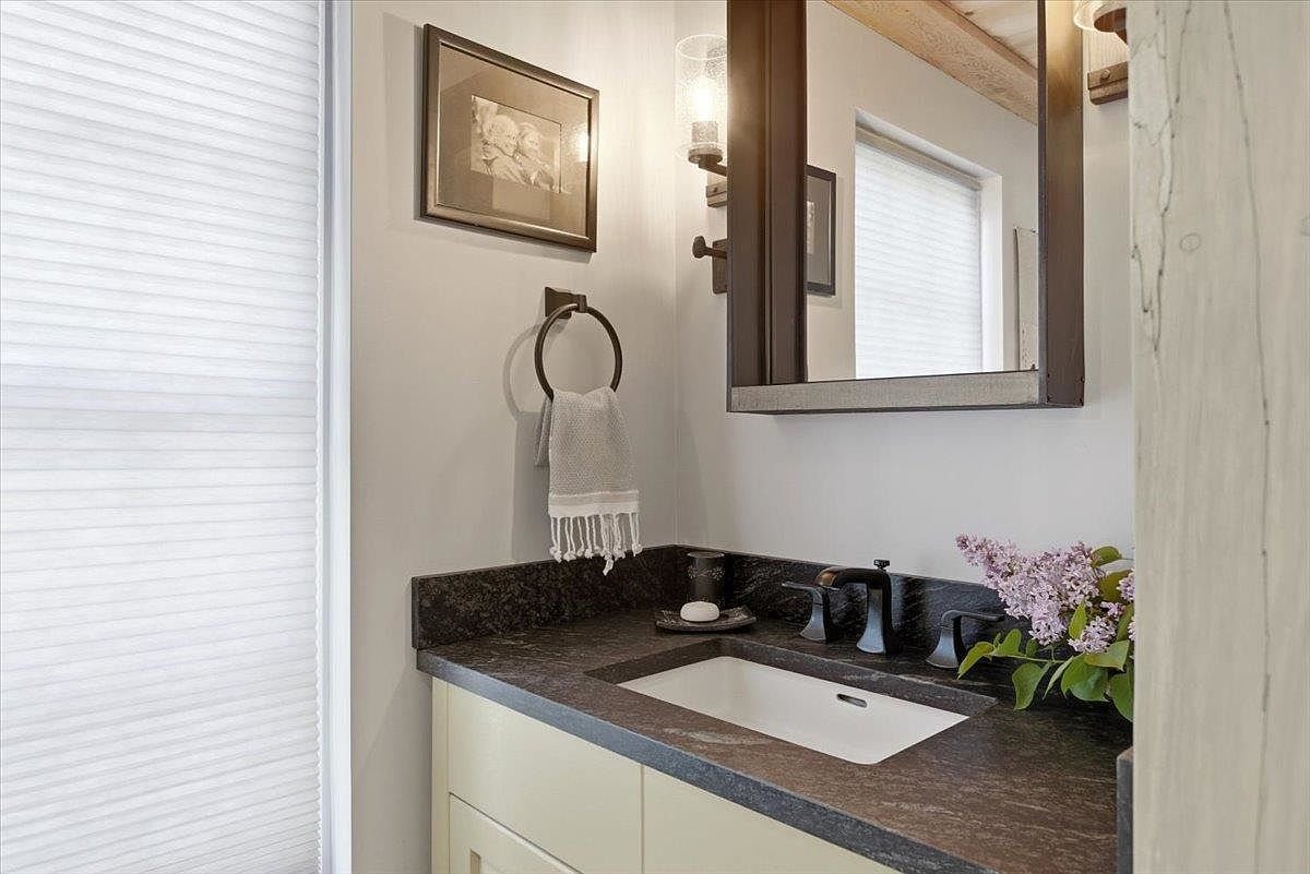 This compact bathroom features a dark, textured stone countertop with an integrated rectangular sink and dark metal fixtures. A framed mirror hangs above the vanity, complemented by a wall-mounted light fixture and a framed picture on the adjacent wall. A small towel ring holds a textured hand towel, and a vase of purple flowers adds a touch of color to the neutral, modern space.