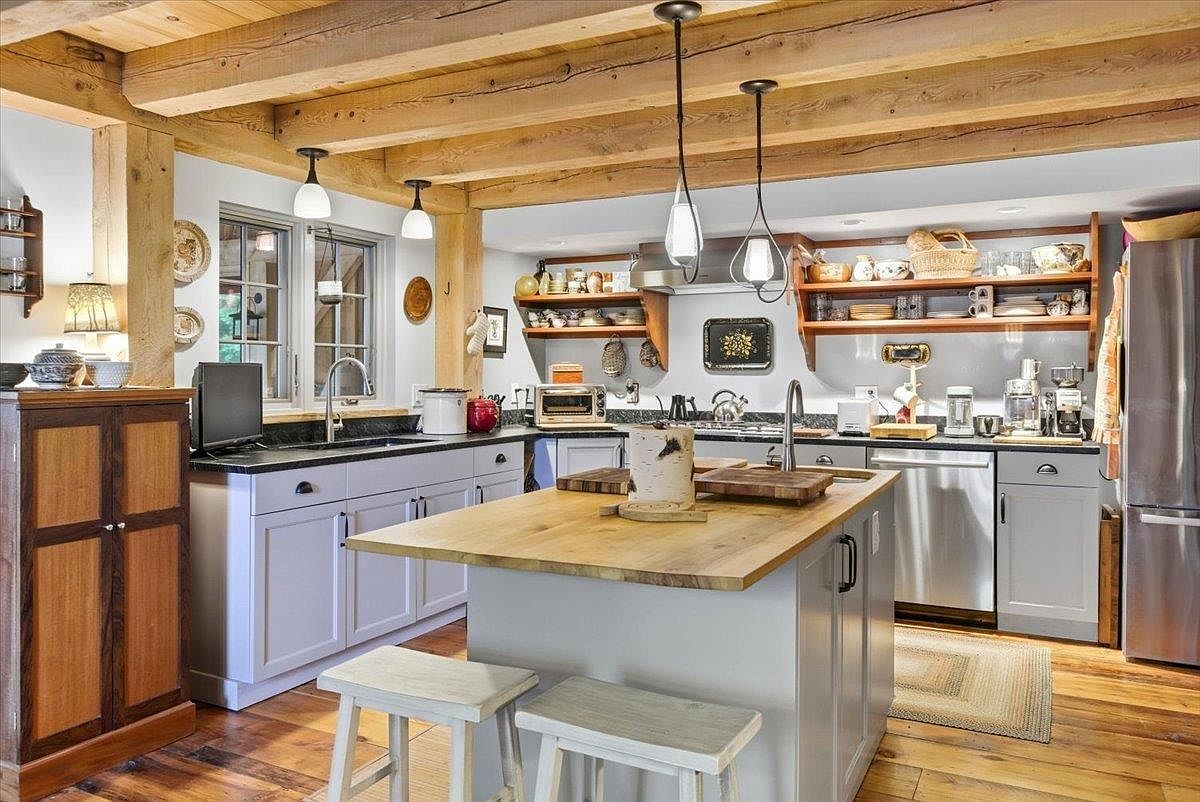 This rustic-inspired kitchen features exposed wooden ceiling beams and warm wood flooring, creating a cozy and inviting atmosphere. The space is equipped with light-colored cabinetry, a central island with a wood-topped counter, and stainless steel appliances. Pendant lights hang above the island, while open shelving provides a charming display area for kitchenware.