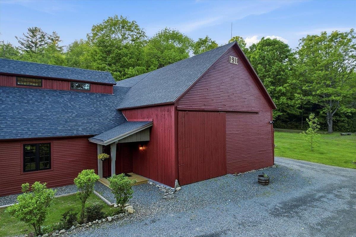 This image captures the exterior of a charming, red-sided barn-style home featuring a prominent '1911' date plaque on the gable. The structure includes a covered entryway with a small porch and a large sliding barn door, set against a backdrop of lush green trees and a gravel driveway. The perspective is slightly elevated, showcasing the architectural blend of rustic barn aesthetics with modern residential updates.