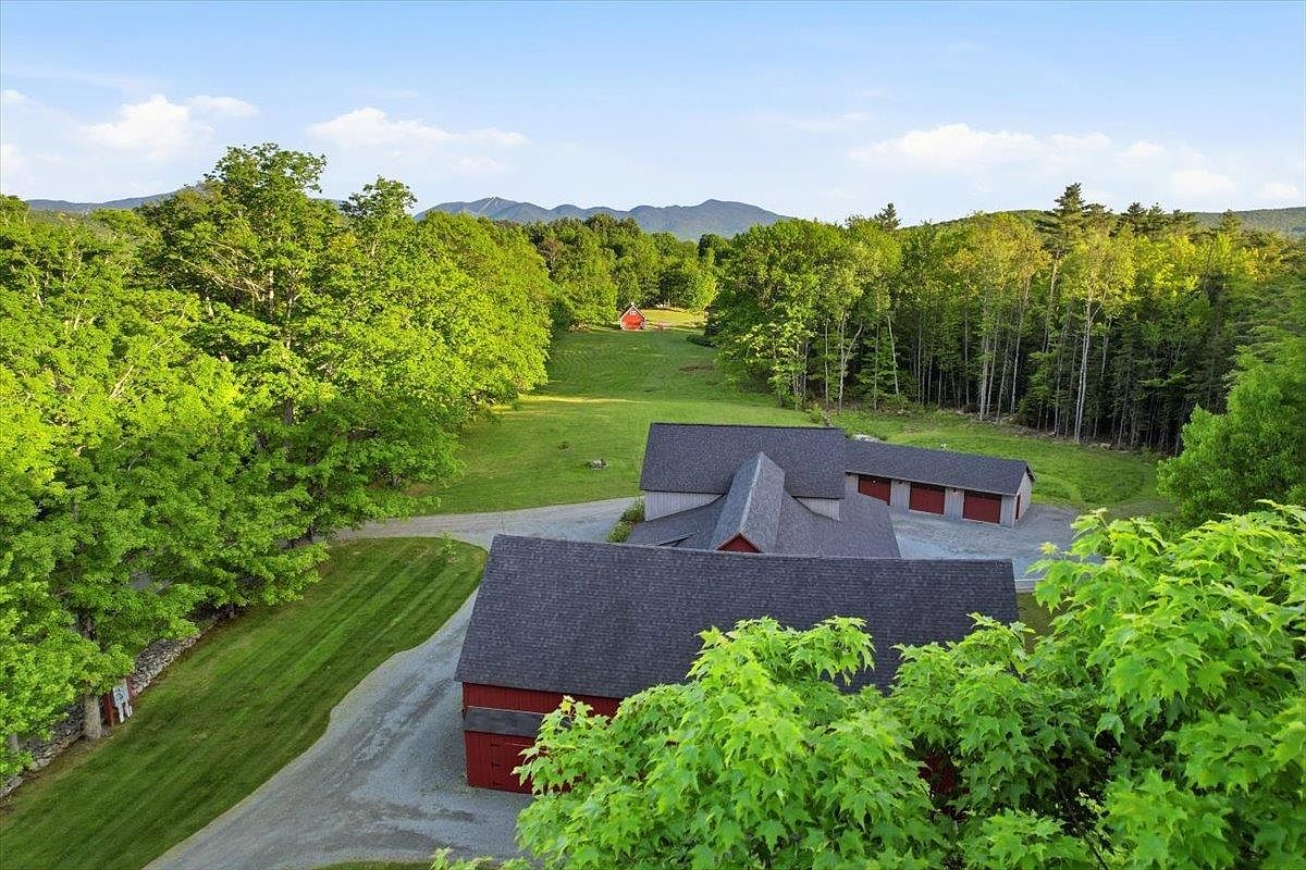 This high-angle aerial view captures a sprawling rural property featuring a large red barn-style structure with a dark shingled roof and an attached garage wing. A long, manicured lawn stretches from the main buildings toward a distant, smaller red outbuilding nestled against a backdrop of dense forest and rolling mountains. The scene conveys a sense of expansive privacy and classic countryside charm, highlighted by the vibrant green landscape under a clear blue sky.