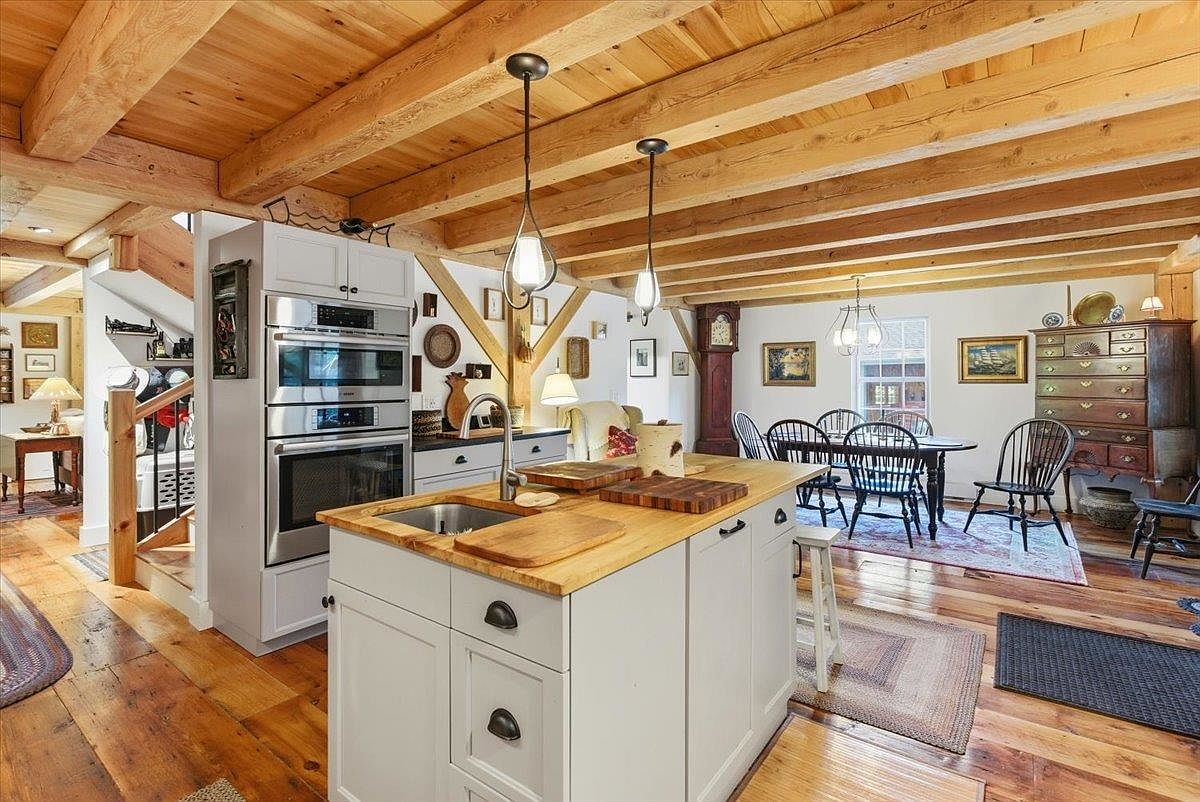 This rustic-style kitchen features exposed wooden ceiling beams and warm wood flooring, creating a cozy and inviting atmosphere. A central island with a light wood countertop and a built-in sink serves as the focal point, complemented by white cabinetry and stainless steel double wall ovens. The open-concept layout flows seamlessly into a dining area, showcasing a blend of traditional charm and functional design.