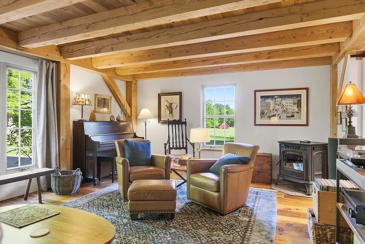 This cozy living room features exposed timber ceiling beams and warm wood flooring, creating a rustic and inviting atmosphere. The space is furnished with two leather armchairs, a matching ottoman, and a piano, all arranged on a patterned area rug. A wood-burning stove sits in the corner, while large windows provide natural light and views of the surrounding landscape.