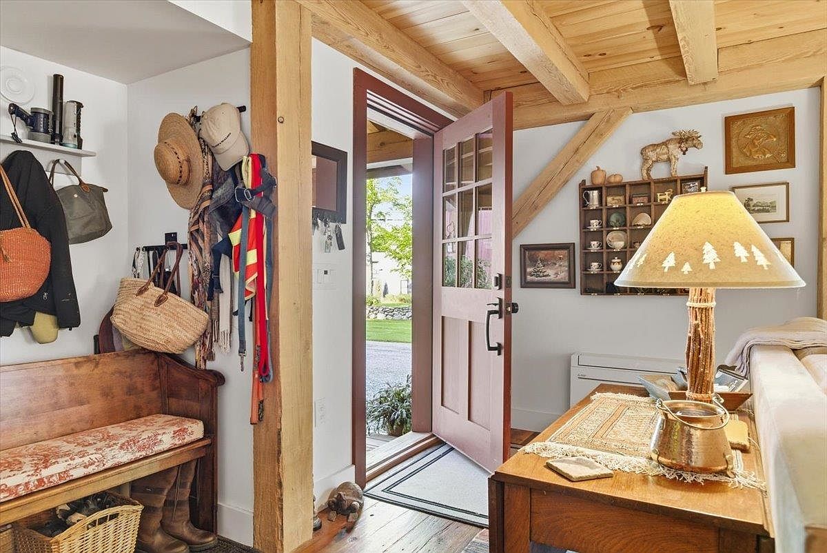 This rustic entryway features exposed wooden beams and warm wood flooring, creating a cozy, cabin-like atmosphere. The space includes a built-in wooden bench with patterned cushions, a wall-mounted cubby shelf for decor, and a lamp with a nature-themed shade resting on a wooden console table. An open door reveals a glimpse of the outdoor greenery, inviting natural light into the welcoming foyer.