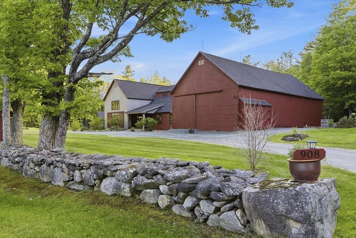 This image captures a charming, rustic-style property featuring a large red barn-style structure connected to a multi-level home with light-colored siding. A classic dry-stacked stone wall runs along the foreground, leading the eye toward the gravel driveway and the lush green lawn. The scene is framed by mature trees under a clear blue sky, creating a peaceful and picturesque rural setting.