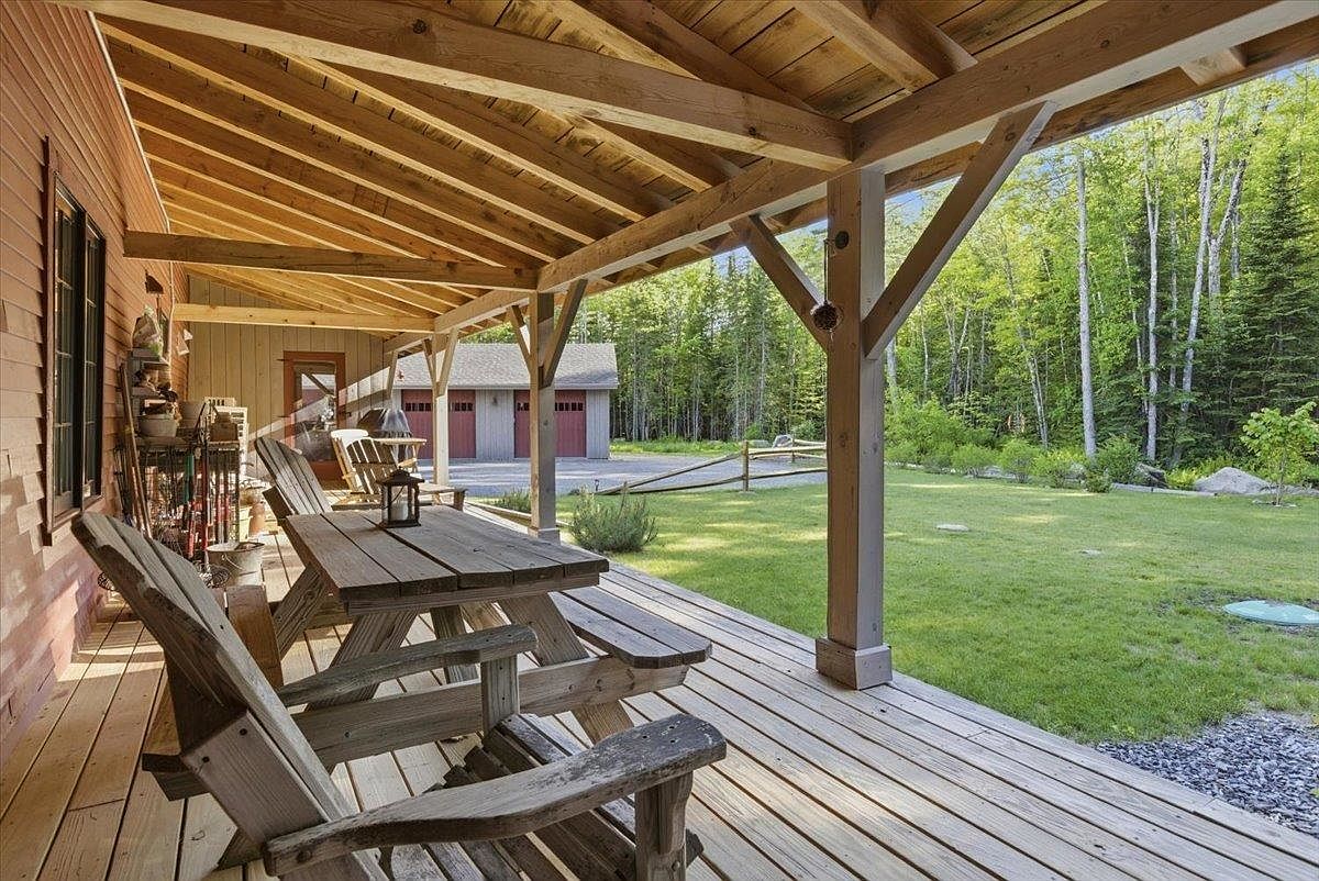 This image captures a spacious, covered wooden deck featuring rustic outdoor furniture, including a picnic table and chairs. The structure showcases exposed timber framing and a natural wood aesthetic, providing a comfortable transition between the home and the expansive, grassy backyard. Beyond the deck, a detached garage with red doors is visible, set against a backdrop of lush, mature trees.