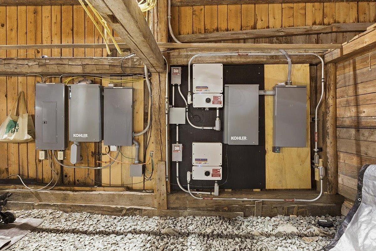 This image shows a utility area inside a rustic structure, likely a barn or basement, featuring exposed timber walls and a gravel floor. A complex array of electrical panels, including two units branded with the Kohler logo, is mounted on the wooden wall and a black backing board, with metal conduit piping running throughout. The scene highlights the functional, industrial infrastructure of the property, emphasizing the mechanical systems in place.
