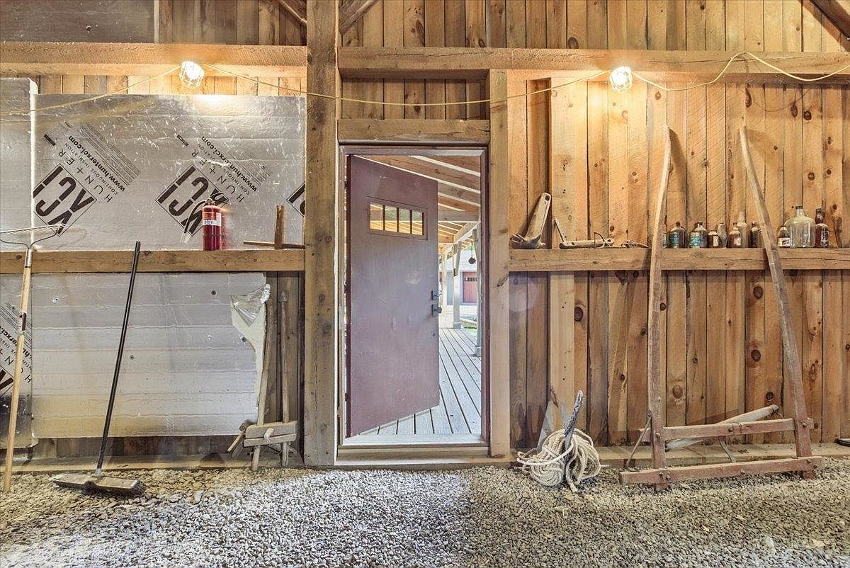 This image shows the interior of a rustic, unfinished barn or storage space with exposed wood plank walls and a gravel floor. An open doorway in the center leads to an exterior wooden deck, while the walls are adorned with various vintage tools, glass bottles, and insulation panels. The scene captures a raw, utilitarian aesthetic with warm, ambient lighting provided by string lights.