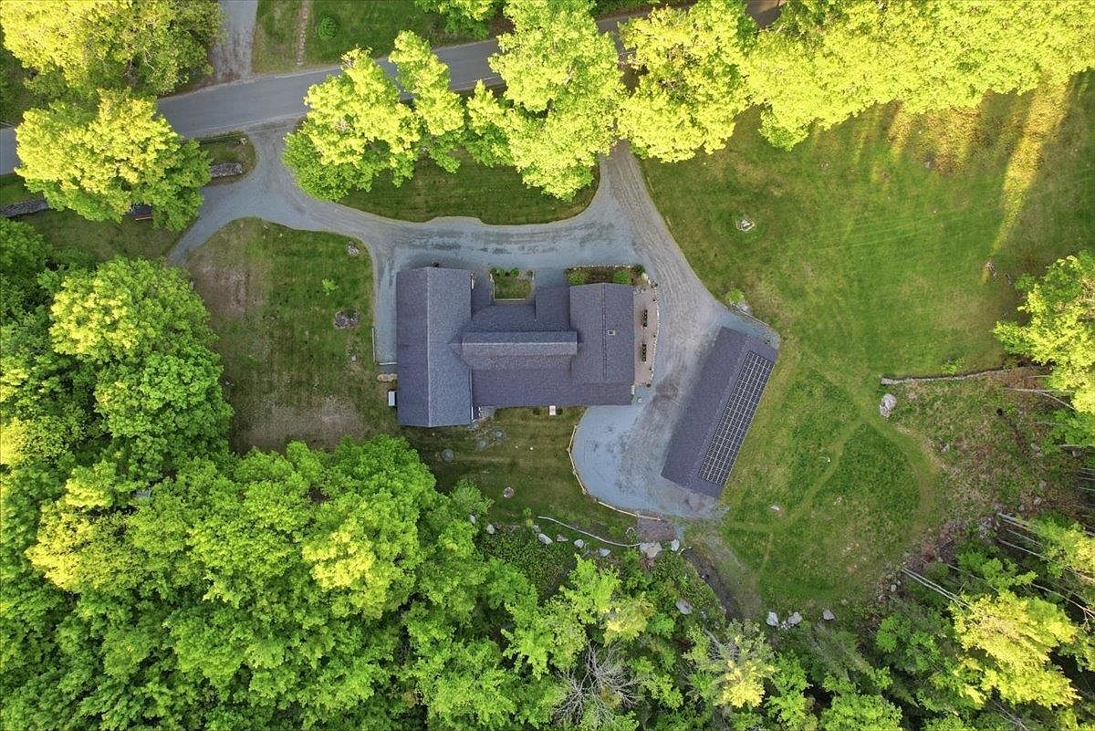 This high-angle aerial perspective captures a residential property nestled within a lush, wooded landscape. The home features a multi-gabled roofline and is accompanied by a separate detached garage equipped with solar panels, all connected by a winding gravel driveway. The surrounding grounds are a mix of manicured lawn and dense forest, providing a secluded and serene setting.
