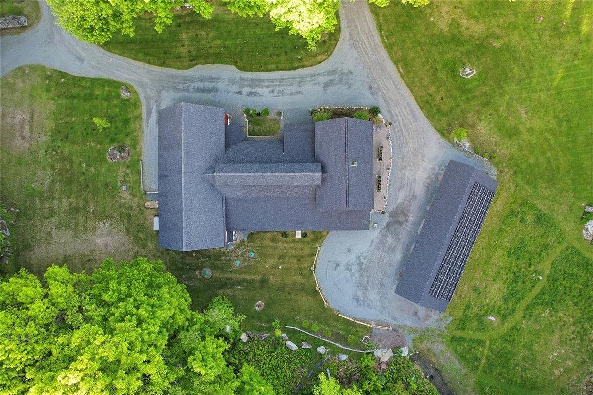 This top-down aerial perspective captures a residential property nestled within a lush, green landscape. The main house features a complex, multi-gabled roofline, while a separate detached structure to the right is equipped with a large array of solar panels. A gravel driveway winds through the property, connecting the buildings and providing access to the surrounding lawn and mature trees.