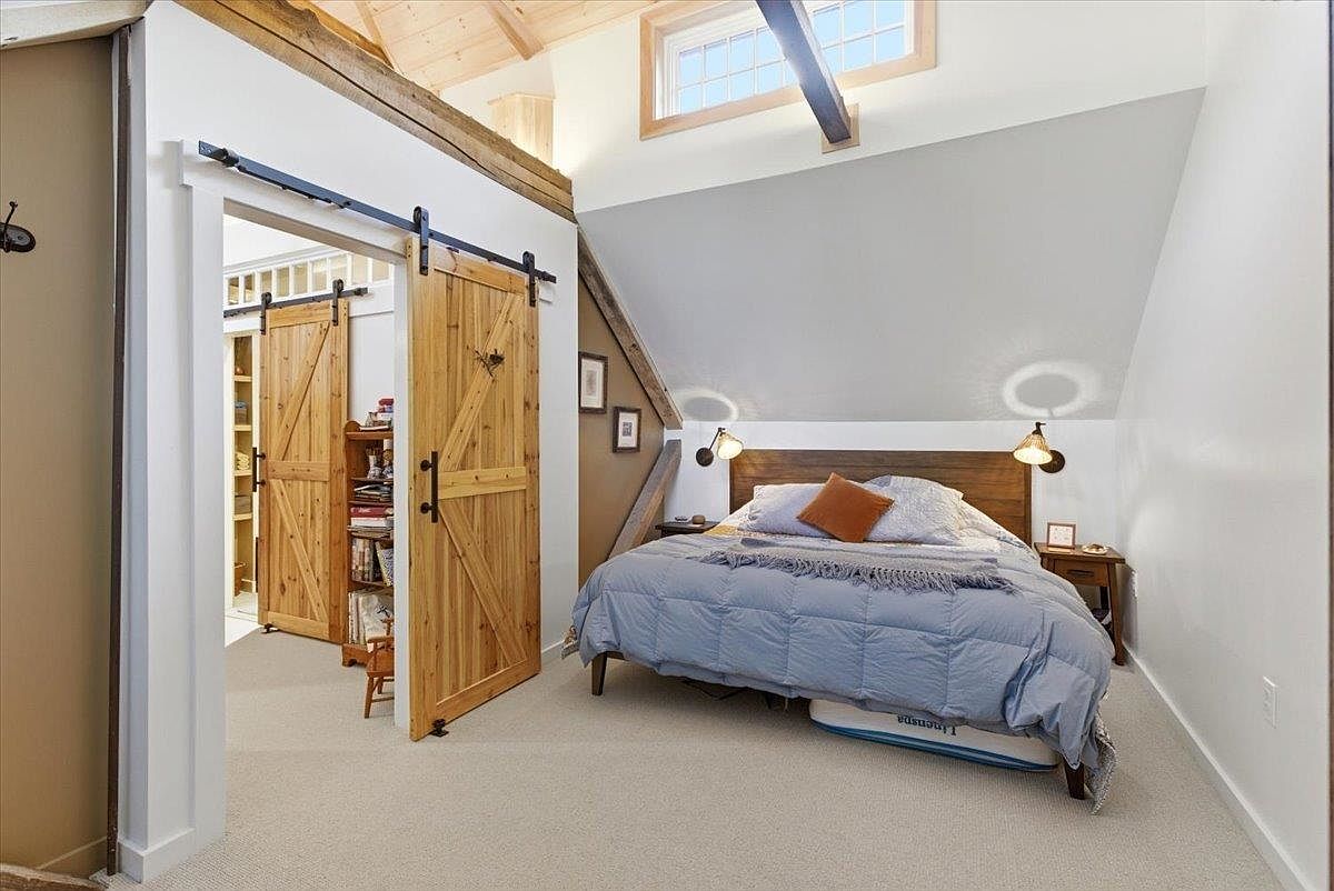 This cozy bedroom features a sloped ceiling with exposed wooden beams and a skylight, creating a warm, rustic atmosphere. A queen-sized bed with a blue comforter sits against a dark wood headboard, flanked by two small bedside tables with wall-mounted lamps. To the left, a sliding barn door opens into a walk-in closet or storage area, adding a touch of farmhouse charm to the space.