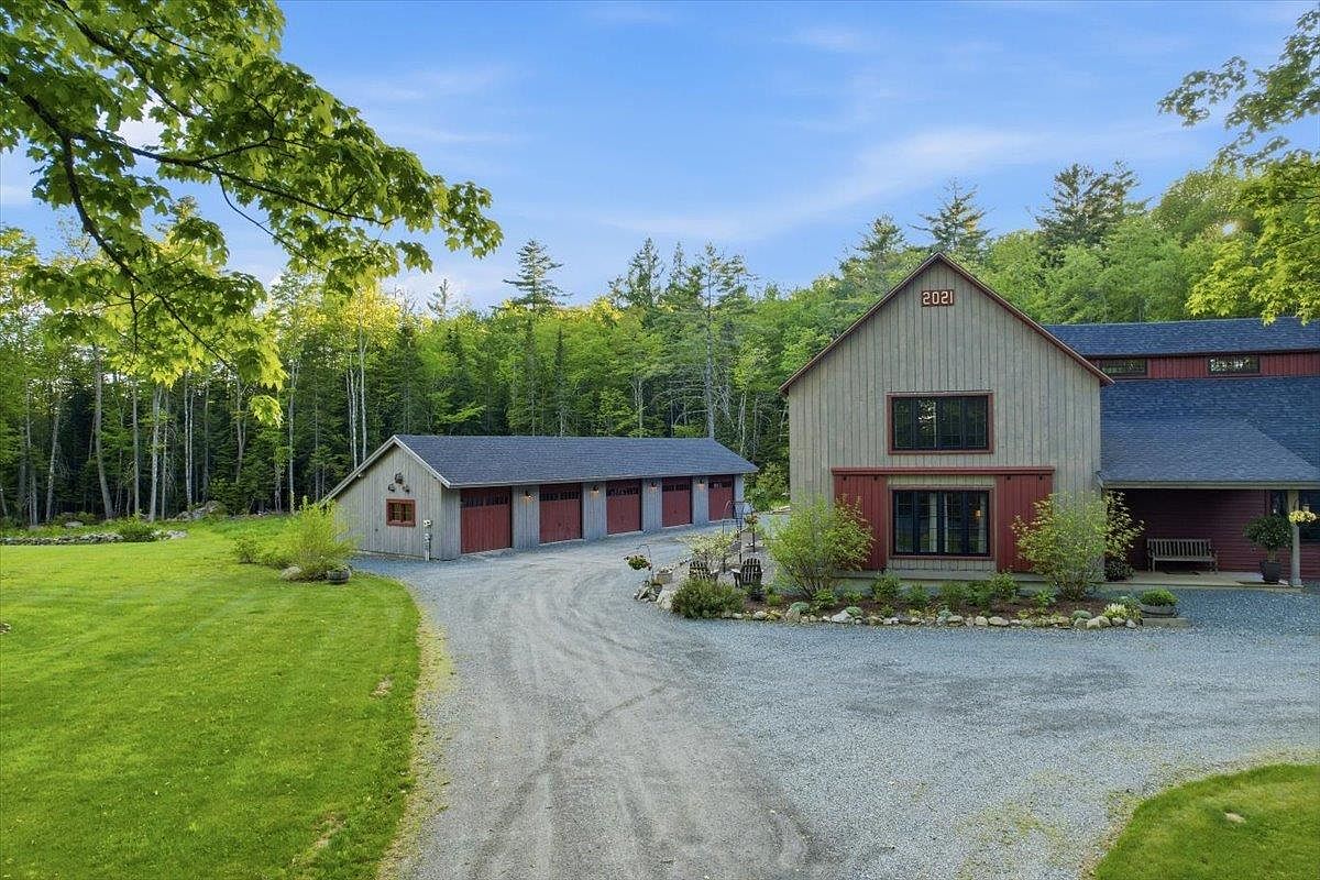 This exterior view showcases a modern farmhouse-style home with a detached multi-bay garage, set against a backdrop of lush green forest. The main structure features vertical siding, dark-framed windows, and a prominent '2021' date plaque on the gable, while a gravel driveway connects the two buildings. The scene is framed by a vibrant green lawn and mature trees, creating a serene and well-maintained rural atmosphere.
