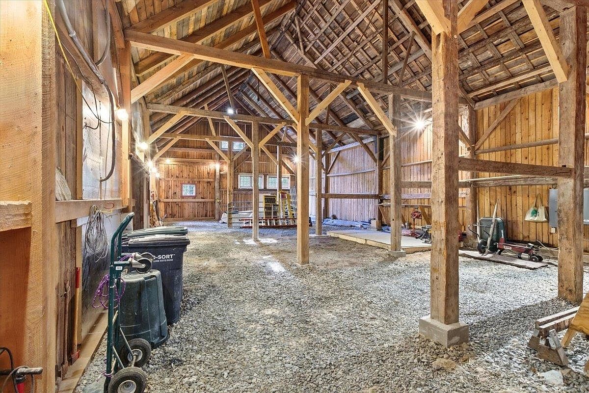 This image captures the interior of a rustic, expansive barn featuring exposed timber framing and a gravel floor. The structure showcases traditional post-and-beam construction with a high, vaulted ceiling and visible rafters, creating a spacious and open atmosphere. The perspective is from the ground level, looking down the length of the barn, highlighting the raw, unfinished aesthetic suitable for storage, workshop, or potential renovation projects.