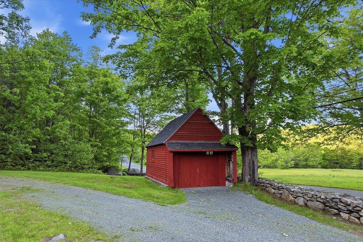 This image captures a charming, rustic red detached garage nestled under the canopy of a large, mature tree. The structure features classic wood siding, a dark shingled roof, and a gravel driveway leading up to its entrance. A traditional stone wall runs along the side of the property, bordering a lush green field and creating a picturesque, rural setting.