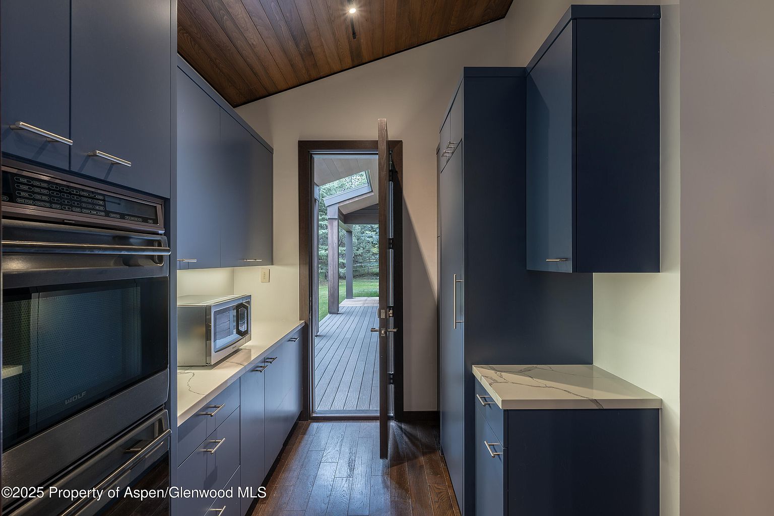 This is a modern kitchen with sleek, dark blue cabinetry and white countertops. Stainless steel appliances, including a built-in oven and microwave, add a touch of sophistication. An open doorway leads to an outdoor deck, providing a glimpse of the surrounding greenery and creating a seamless indoor-outdoor flow.