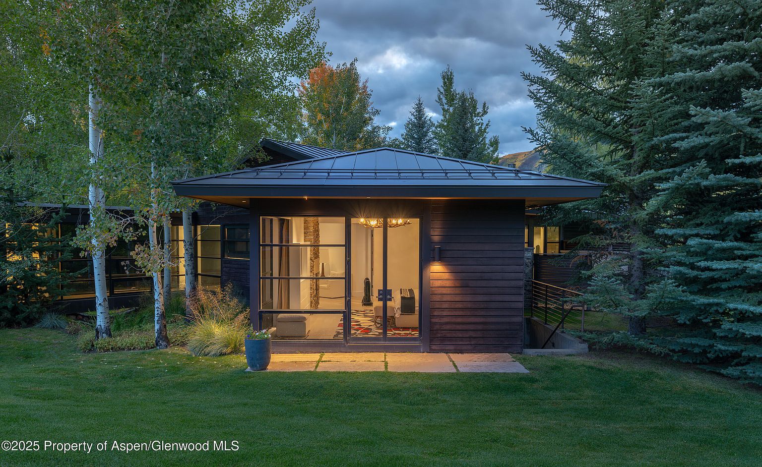 The image showcases the front exterior of a modern home with dark wood siding and a sleek, low-pitched roof. Large windows offer a glimpse into the interior, revealing a chandelier and contemporary furnishings. The property is surrounded by lush greenery, including mature trees and a well-maintained lawn, creating a serene and private setting.