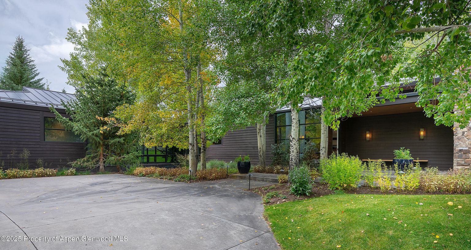 This is a front exterior view of a modern home with dark wood siding and a metal roof. The property features mature trees, landscaping, and a concrete driveway. The entryway has a covered porch with stone accents and outdoor lighting, creating an inviting atmosphere.