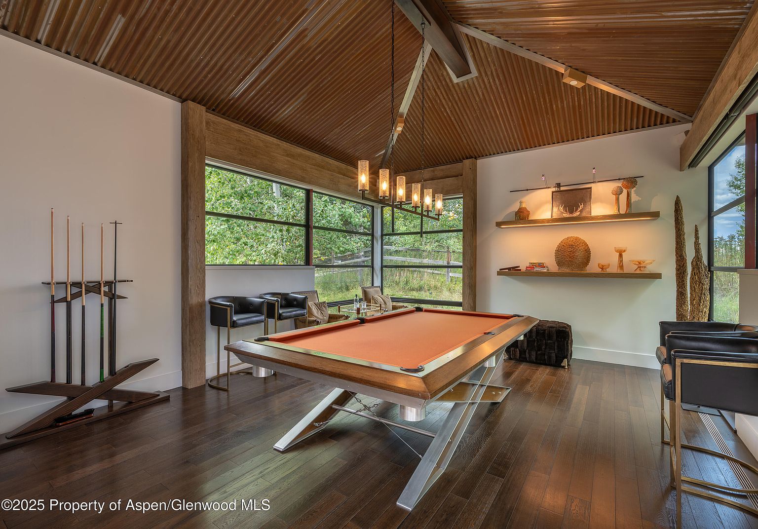 This interior shot showcases a stylish living room featuring a unique pool table as a focal point. The room is well-lit with natural light streaming through large windows, complemented by modern pendant lighting. The dark wood flooring and corrugated metal ceiling add a rustic yet contemporary touch, creating an inviting and sophisticated space.