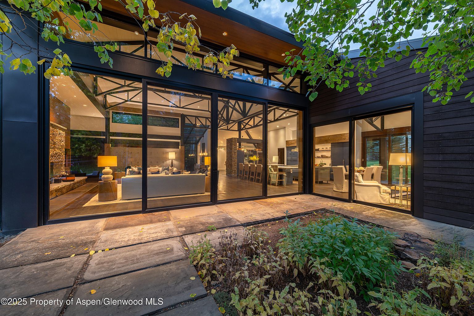 This image showcases a modern home's patio area, seamlessly blending indoor and outdoor living. Large glass doors provide expansive views into the living room, dining area, and kitchen, while a stone patio and garden create an inviting outdoor space. The architectural design emphasizes clean lines and natural materials, creating a sophisticated and tranquil atmosphere.