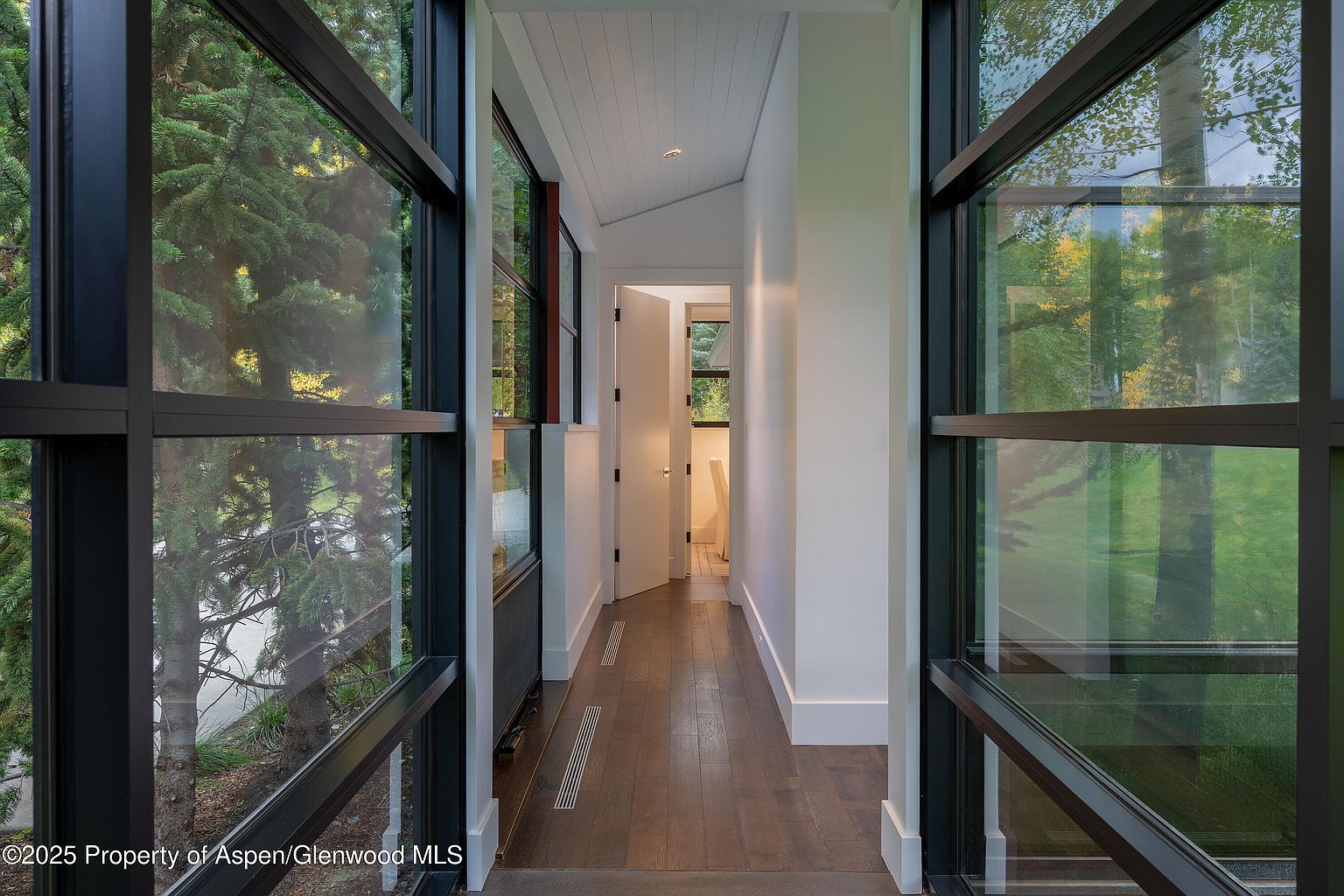 This interior shot showcases a modern hallway with dark wood flooring and white walls, framed by large windows with black frames offering views of the surrounding greenery. The hallway leads to a partially open white door, suggesting access to another room. The design emphasizes natural light and a seamless connection with the outdoors, creating a bright and airy atmosphere.